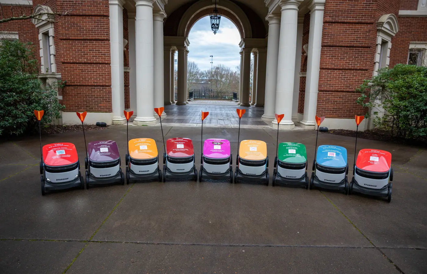 A mix of different colored delivery robots in a row in front of Weatherford Hall.