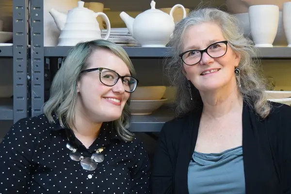 Two people pose and smile in front of pottery on a shelf.